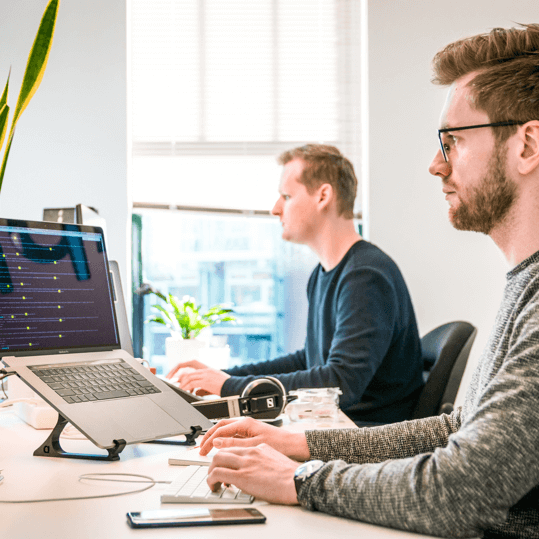 Two men working at desks in a modern office, one using a laptop on a stand and the other focused on a desktop computer.