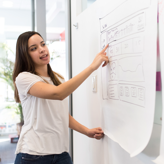 Woman in a white shirt presenting data and charts drawn on a large paper sheet attached to the wall.
